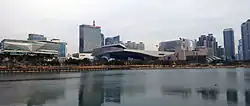 Photo couleur d'un ensemble de bâtiments, dont plusieurs gratte-ciel. Un cours d'eau au premier plan, un ciel nuageux en arrière-plan.