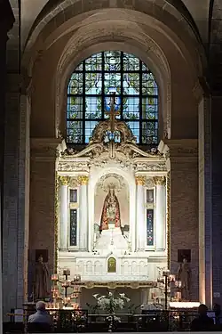 Photographie de la chapelle de Notre-Dame des Miracles et des Vertus. La statue de la Vierge à l'Enfant, l'un et l'autre couronnés, drapée dans un manteau rouge, est placée au centre d'un retable en marbre blanc. Un vitrail emplit la baie au-dessus du retable. Des fleurs et de nombreux cierges se trouvent devant l'autel. Les murs de la chapelle sont couverts d'ex-voto. Deux fidèles prient devant l'autel.