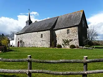 Bâtiment massif en pierre grise et toit d'ardoise noire, au milieu d'une étendue herbeuse tondue ras. On aperçoit le toit du clocher à l'arrière. Au premier plan, une clôture en bois