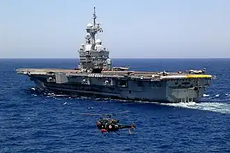 Photographie couleur montrant le porte-avion français Charles de Gaulle en cours de catapultage d'avions super-étendard. La vue de trois quart arrière à gauche, permet de voir l'alouette III en premier plan qui surveille les envols.