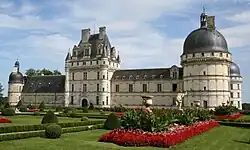 Photographie d'un imposant château à pierres blanches, présentant une tour centrale carrée et à toit droit, une aile droite terminée par une large tour ronde à toit en dôme, une aile gauche plus petite, terminée par une petite tour ronde. Au premier plan, un jardin à la française : herbe tondue, buis et parterre de fleurs.