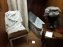 Photographie d'une vitrine du musée. Sur la gauche, sur une chaise, est exposée une chemise française du XVIIIe&nbsp;siècle à col échancré orné de dentelles. À droite, sur une petite commode, une perruque grise de la même époque est disposée sur son support. Au fond, on peut voir une baignoire en fer blanc à demi masquée par un rideau clair.