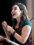 Femme brune souriante et portant un haut vert et des boucles d'oreilles. Elle tient un feutre dans les mains.