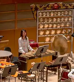 photo de la section percussion d'un orchestre avec une femme jouant d'un instrument au centre et derrière elle, à droite, un yunlao à 37 gongs.