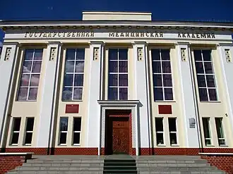 Photographie en contre-plongée d'un grand bâtiment jaune avec de grandes fenêtres et de grandes colonnes. Au-dessus des colonnes est inscrit en russe « Académie médicale d'État »