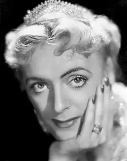 Femme aux cheveux courtes, portant une couronne et tenant sa tête dans sa main. Elle porte du vernis à ongle rouge.