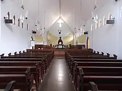 Photographie couleur de l'intérieur d'une église sans bas-côté ni transept, entièrement peinte en blanc, avec des bancs de bois.