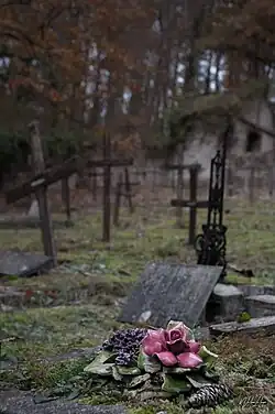 Vue du cimetière des Fous d'Évreux. En avant plan se trouve un objet funéraire représentant un bouquet de fleurs émaillées posé sur une sépulture. En arrière plan se trouve des croix de bois et le mur d'enceinte du cimetière ainsi qu'un petit bâtiment dégradé.