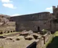 Photographe de ruines d'une infrastructure romaine en brique, partiellement ensevelie sous la végétation, avec un mur long et imposant en arrière-plan.
