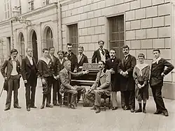 Vue en noir et blanc de 11 hommes et une femme debout derrière un piano devant lequel sont assis 2 hommes.