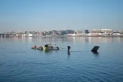Photo de l'épave de l'hélicoptère. Un morceau de carlingue en partie arraché, une antenne, une roue et la dérive sont partiellement émergés. On distingue l'aéroport en arrière plan, au bord du fleuve.