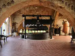 la photographie en coleur représente une pièce du musée codorniu. Une voute au premier-plan sert de support à des outils viticoles : fourche-bèches et sarclettes. Au second plan, un pressoir vertical ancien à cage en bois occupe le centre de la pièce. Des outils anciens, un mini pressoir ou une amphore en terre cuite occupent les angles de la salle.