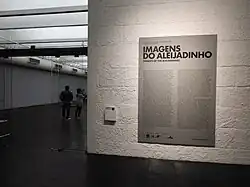 Photographie en couleurs. Entrée d'une exposition, où un mur affiche le texte explicatif de l'exposition.