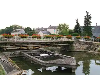 Photo couleurs. Le pont tournant de Combleux est vu de côté. Il est constitué d’une poutre métallique ajourée reposant sur une articulation. Au droit de celle-ci on devine l’emplacement du mécanisme faisant pivoter l’ouvrage. Devant se situe une sorte d’estacade. Les garde-corps du pont portent des pots de fleurs rouges.