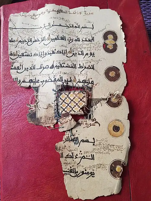 Description d'image : photographie représentant un fragment ancien d'un manuscrit coranique annoté avec l'alphabet arabe en vieux-kanembu, posé sur un support de cuir rouge foncé. Le manuscrit est décoré de plusieurs motifs circulaires en brun et jaune sur les marges, ainsi qu'un motif de dessins géométriques au centre du manuscrit. Le motif tient dans le manuscrit grầce à du fil cousu à travers le papier. La couture est relâchée, laissant voir le support, distinguant le motif du parchemin. De plus, le parchemin montre des traces d’usure et des réparations anciennes avec du fil cousu à travers le papier.