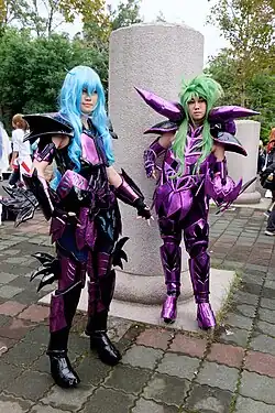 Deux personnes déguisées, portant des armures violettes sophistiquées, ainsi que des perruques vertes et bleues.