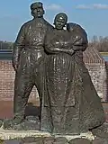 Photographie en couleurs d'une sculpture montée sur un socle en pierre et d'une voie d'eau en arrière-plan