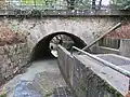 photo d’un pont sous lequel passe une eau boueuse ; au milieu du passage, dans le sens de l’eau, se trouve un mur ; la partie droite est normalement un passage pour les piétons