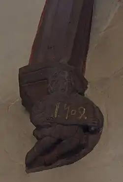 photographie en couleur montrant un culot en grès rose sculpté avec un personnage barbu et vêtu d’habits médiévaux, qui tient une banderole inscrite avec la date 1504