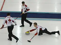 Photographie de trois curleurs, l'un à droite en position allongée, genou plié, tenant dans la main droite une pierre, dans la main gauche un balai, les deux autres debout portant un balai.
