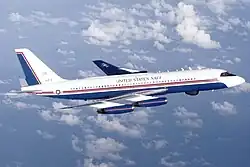 Photo couleur d'un avion en vol, vu du côté droit. Aux couleurs blanches, bleues sur le ventre et disposant de fines bandes rouges au centre, l'avion vole dans un ciel bleu parsemé de quelques nuages.