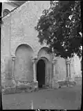 Porte en arc romane en pierre grise, entourée d'arbres, éclairage diffus, atmosphère paisible.