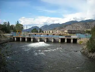 Photo d'un barrage prise depuis un lac, qui semble faire une vingtaine de mètres de large, un portail métallique est visible sous le barrage, une rambarde et un grillage bleu se trouvent au dessus. Derrière le barrage de bâtiments et des montagnes sont visibles.