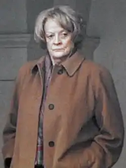 Photo en demi-profil (gauche) d'une femme au visage ridé, qui se tient adossée à une colonne en pierre avec les mains dans les poches. Ses cheveux sont grisonnants, coupés «&nbsp;au bol&nbsp;». Elle porte un trench-coat marron et son regard est concentré sur quelque chose situé à la droite du photographe.
