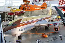 Vue plongeante, de trois-quarts avant, d'un avion de couleur jaune, marron et gris dans le hall d'un musée ; plusieurs autres avion, plus petits, sont exposés tout autour, au sol ou suspendus en l'air.