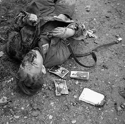 Photo noir et blanc prise le 23 décembre 1943 à Ortona, en Italie. Dans la partie gauche de la photo, le cadavre d'un soldat allemand gît (tête vers le bas de la photo) sur un sol de terre pierreuse. Trois photos de soldats sont visibles près de son épaule droite.