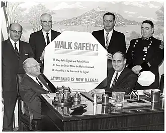 Des hommes politiques entourant un panneau sur lequel est notamment écrit « Walk safely ! Jaywalking is now illegal ».