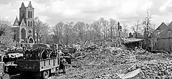 Photographie en noir et blanc montrant les ruines d'une ville, avec les murs d'une église (sans toit) encore visible. Au premier plan, un camion militaire.