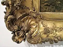 Gros plan d'un cadre photo doré et décoré de sculptures florales complexes et d'un motif de coquillage. Le cadre présente une surface texturée avec un mélange de finitions mates et brillantes, mettant bien en valeur le savoir-faire.
