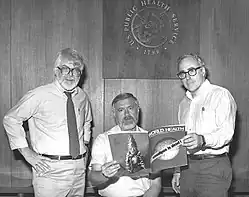 Trois hommes se mettent en scène au Centre pour le contrôle et la prévention des maladies aux États-Unis. À gauche, Dr J. Donald Millar, en cravate et chemise blanche, mains sur les hanches. Au centre, Dr William H. Foege, en t-shirt, assis, tenant dans sa main droite un exemplaire du périodique de l'Organisation Mondiale de la Santé de Mai 1980 montrant en couverture un globe terrestre barré par un message : "Smallpox is dead!" ("La variole est morte !"). À droite, Dr J. Michael Lane, de trois-quart, en chemise blanche et tenant le même exemplaire du périodique avec sa main gauche. Tous regardent la caméra. Sur le mur laqué au dessus d'eux, on aperçoit le logo des services publics de santé États-Uniens (en anglais : "U.S. Public Health Service").