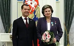 Homme blanc en costume souriant se tenant à gauche d'une femme en costume tenant un bouquet de fleurs