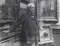 Paul Durand-Ruel debout s'appuie contre un mur de sa galerie où plusieurs tableaux sont accrochés.