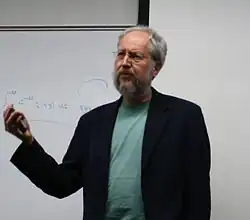Un homme debout devant un tableau d'école, dont ont ne voit que le haut du corps depuis la taille, lève le bras droit.