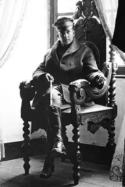 Un homme est assis dans une chaise richement décorée. Il porte un képi, un manteau et des bottes de cavalier et il tient une cravache.