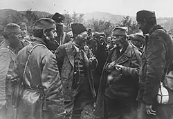 Un homme barbu en uniforme, coiffé d'une toque de fourrure, entouré d'autres hommes en uniforme.