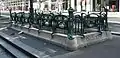 Photo en couleurs d'un entourage à cartouches de fonte ajourés posé sur le trottoir séparant une rue et une contre-allée
