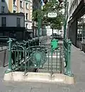Vue en perspective se prolongeant vers le haut d'une bouche de métro, avec contraste entre le gris vert des ferronneries et le vert émeraude intense d'une poubelle placée dans le prolongement des escaliers.