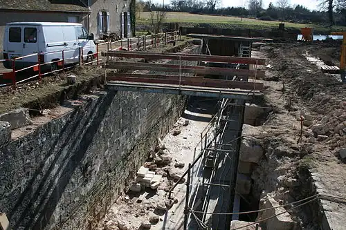  Photo couleurs. L’écluse du bas de Grignon, vue en perspective frontale, est en travaux. Elle est à sec, on peut voir au fond de la photo, les palplanches faisant office d’étanchéité. À droite, le mur de droite de l’écluse (bajoyer) est entièrement démonté pour que l’ensemble des pierres le constituant soit remplacé par des pierres de taille neuves. Sur la partie gauche, seul le couronnement (partie haute du mur) est démonté. Une passerelle métallique de chantier traverse l’écluse. Des pierres de taille neuves reposent ne fond d’écluse.