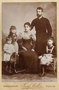 Photo sépia montrant un homme barbu debout, une femme assise en robe longue, une fillette assise, une autre debout, et un garçonnet sur un cheval de bois