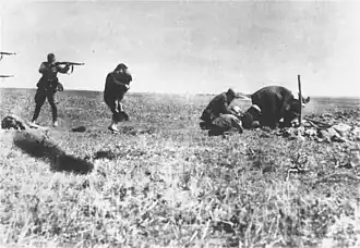 Photo noir et blanc prise à Ivangorod, en Ukraine. Dans un champ herbeux, sous un ciel clair, un soldat allemand (à gauche, au second plan), de profil, jambes écartées, tient en joue une femme qui lui tourne le dos, un enfant dans les bras. Devant le soldat, on distingue un corps de femme étendu sur le sol. Sur le bord gauche de la photo, deux canons de fusils, l'un au-dessus de l'autre, sont aussi visibles. Et, sur la droite, quatre hommes semblent creuser un trou.