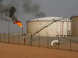Au premier plan, un grillage est installé sur le sable, devant deux bâtiments circulaires contenant des hydrocarbures, à droite. En arrière-plan à gauche se dresse une torchère enflammée.