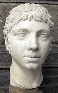 Tête sculptée d'un jeune homme aux cheveux plus longs, au regard ennuyé, le visage rond, de longues rouflaquettes et à la moustache fine.