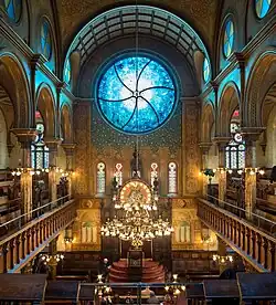 Une grande salle vue de haut. Une grande coupole avec au centre un vitrail bleu. Sur les côtés, des balcons. Vers le bas, un chandelier derrière lequel apparaît le sol.