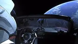 Vue de la Terre depuis l'intérieur d'une voiture. Un écriteau est présent sur le tableau de bord.