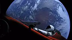 Disque circulaire représentant la Terre entièrement illuminée montrant l’Australie, flottant dans le vide spatial. Devant la Terre, une voiture décapotable rouge vue de côté. Une silhouette humanoïde en combinaison spatiale est assise au volant, le bras droit sur le volant, le bras gauche posé sur la portière.