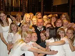 Photographie en couleur d'un homme aux bras écartés, entouré de vingt quatre jeune filles, presque toutes blondes et souriantes sauf une à l'arrière plan, se cachant le visage